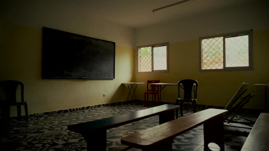 A static, wide shot shows a dimly lit and abandoned classroom with a blank chalkboard and simple wooden benches