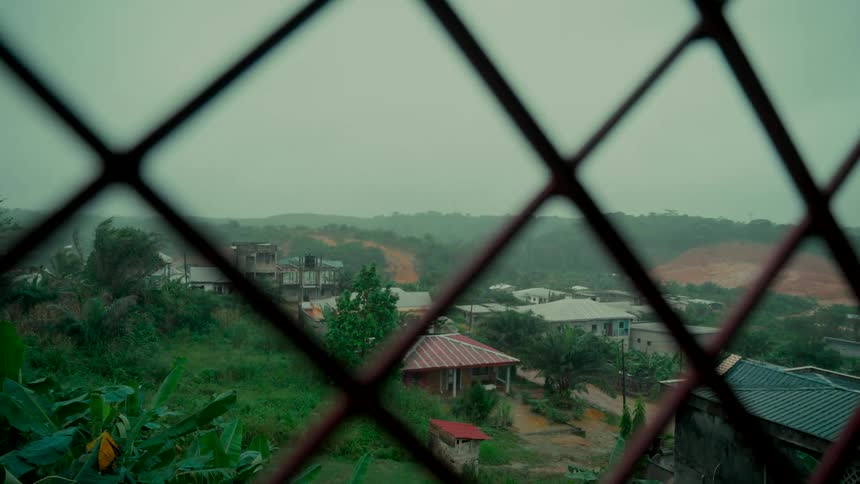 View of a lush green hillside village from behind a grated window during a windy rainstorm