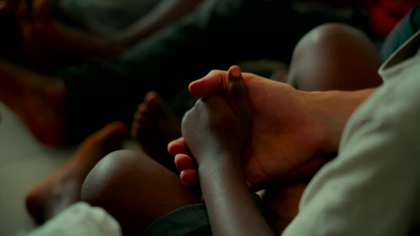 A close-up shot shows a Caucasian adult's hand gently holding the hand of an African child, symbolizing comfort, unity, and support
