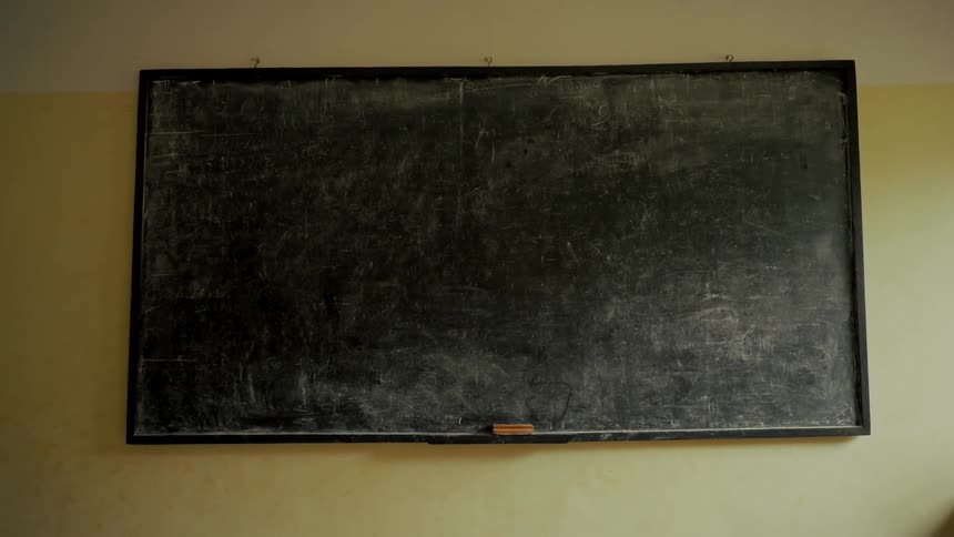 A static medium shot of a blank, smudged chalkboard hanging on a classroom wall with an eraser on the chalk rail