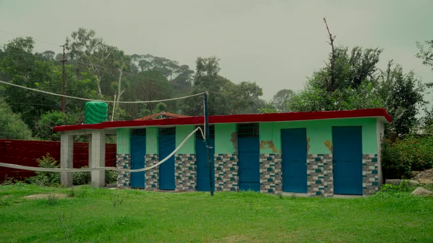 A still shot of a public restroom building with green walls and blue doors in a grassy field surrounded by trees on an overcast day