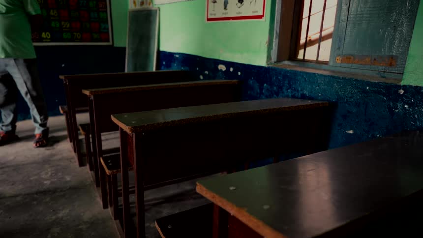 A static shot captures the interior of an old, empty classroom with rows of wooden desks and a teacher standing in the background