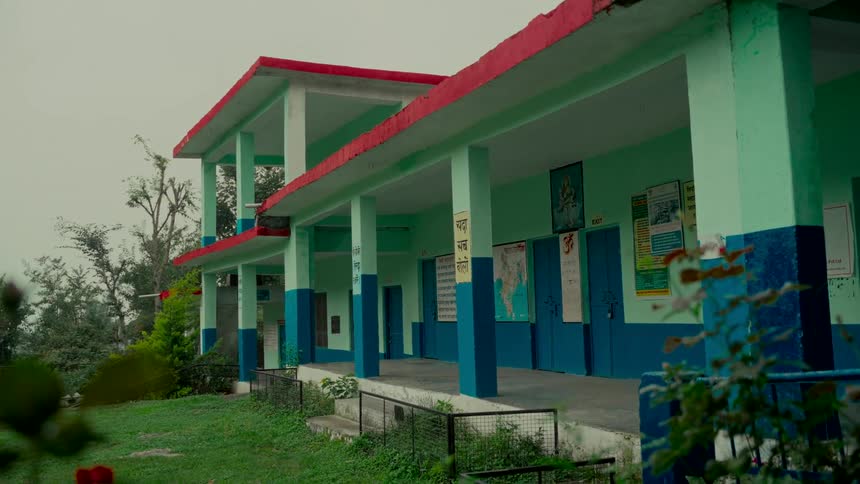 A brightly colored rural school building sits quietly on a green hillside on an overcast day