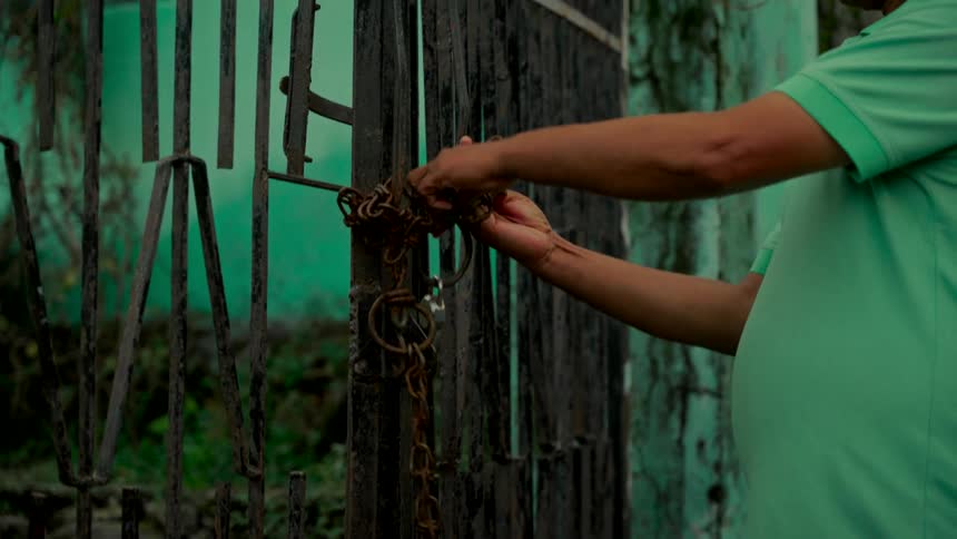 A man in a green shirt struggles to untangle a rusty chain and remove a padlock from an old metal gate