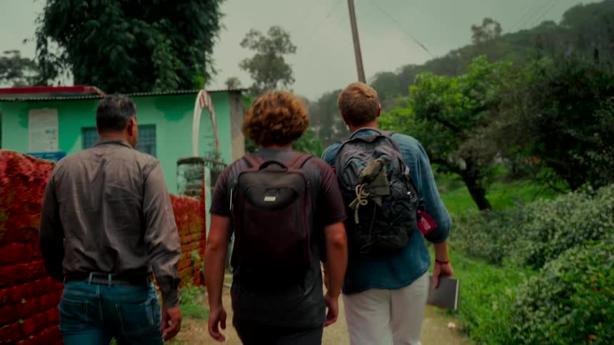 Three men wearing backpacks walk in slow motion down a rural path in a lush, green village