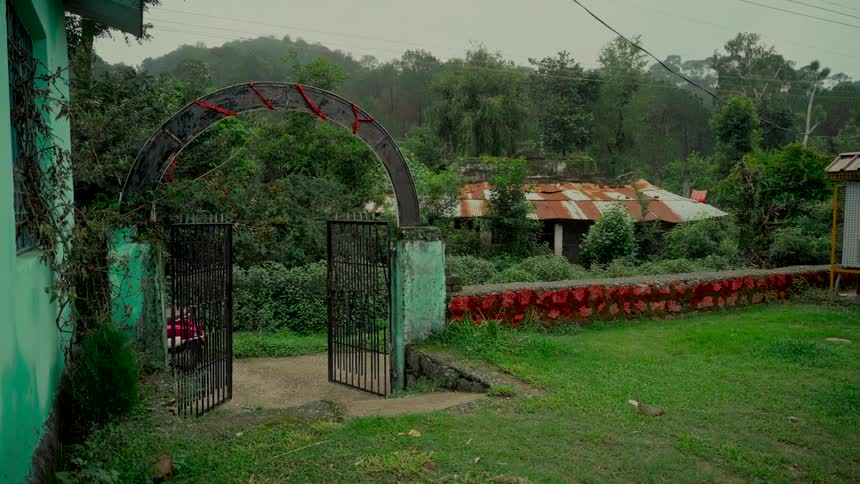 A slow zoom out from an open gate reveals an overgrown, abandoned house with a rusty roof on a gloomy day