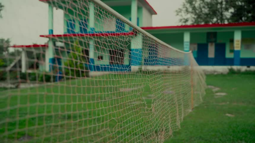A weathered badminton net with holes stands on a green lawn in front of a colorful rural school building