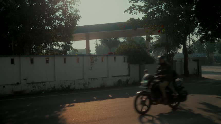 A person on a motorcycle drives in slow motion past a gas station on a hazy, sunlit street