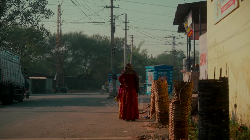 A woman in a traditional red dress waits patiently on the side of a hazy, sunlit road in a small Indian town