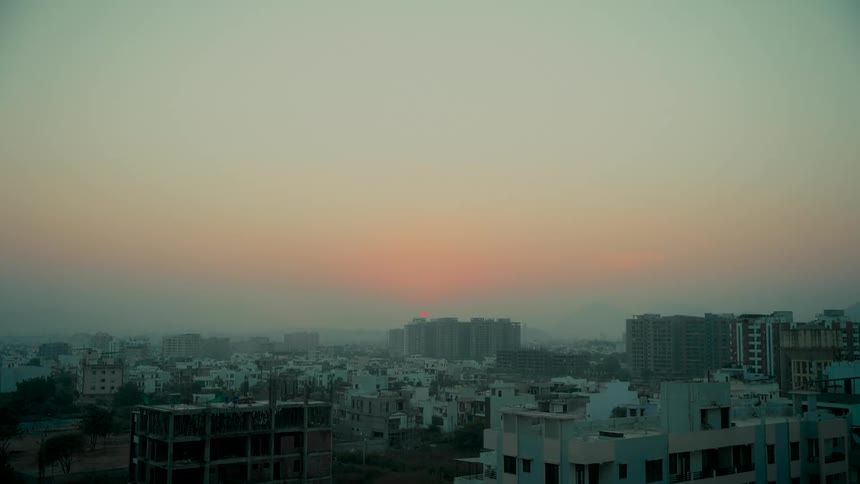 The sun sets behind the buildings of a hazy city skyline during a calm twilight
