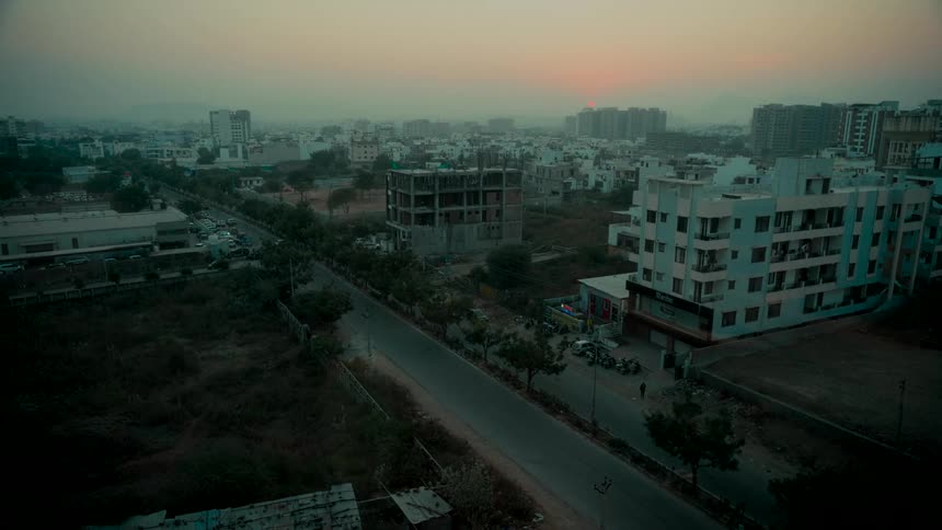 A wide, static shot of a hazy cityscape at sunrise with cars and an auto-rickshaw driving on the street