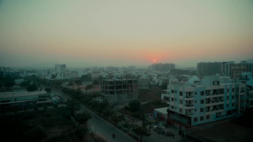 The sun sets over a hazy, sprawling cityscape with a mix of buildings and a quiet street