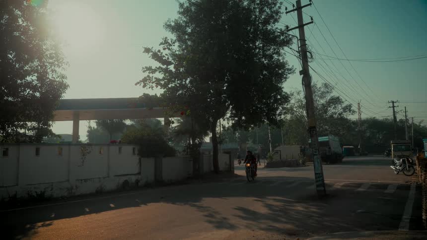 Backlit slow-motion view of a motorcyclist navigating a quiet intersection on a sunny day in India