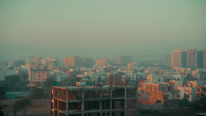 Wide shot of a developing city skyline during a hazy sunrise, with a prominent building under construction in the foreground