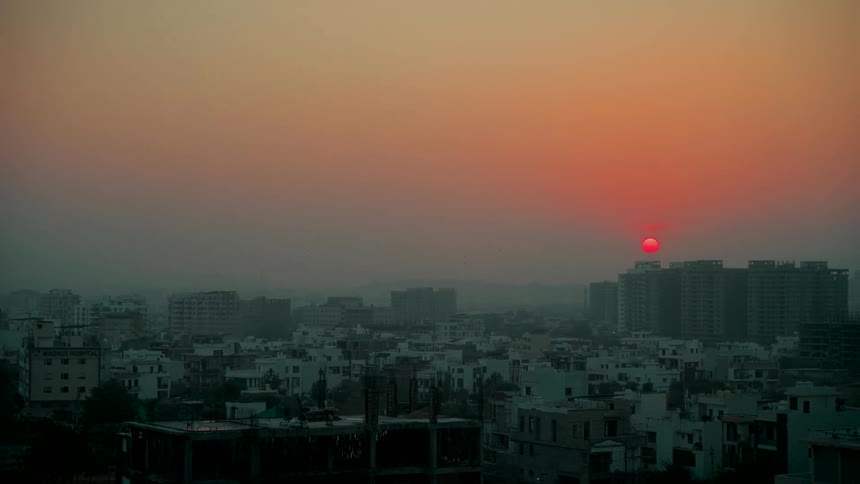 A slow pan reveals a hazy cityscape with a vibrant red sun setting behind buildings at dusk