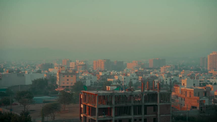 A slow pan across a hazy city skyline in India at sunrise, revealing modern high-rises, residential homes, and buildings under construction