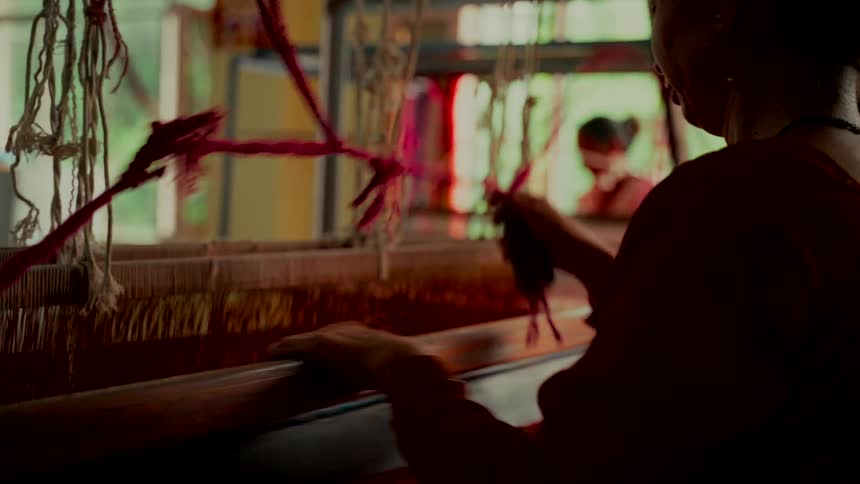 A silhouetted artisan skillfully works at a traditional handloom, weaving colorful threads together in a workshop