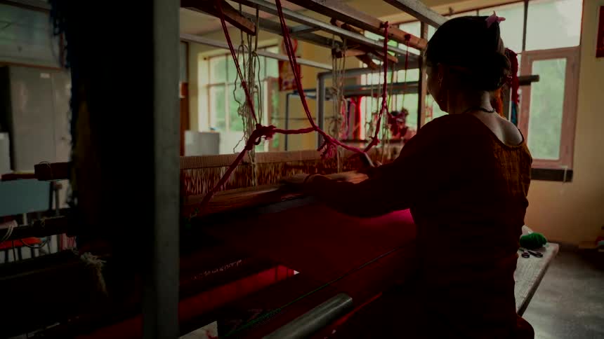 A silhouetted woman works meticulously at a traditional handloom, weaving vibrant red fabric in a sunlit workshop