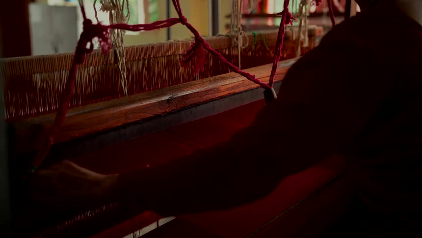 A silhouetted artisan meticulously works a traditional wooden handloom, weaving a vibrant red fabric in a workshop