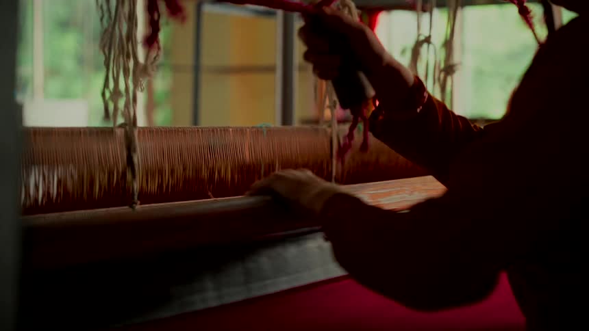 Close-up of a person weaving bright pink cloth on a traditional wooden loom in a dimly lit workshop