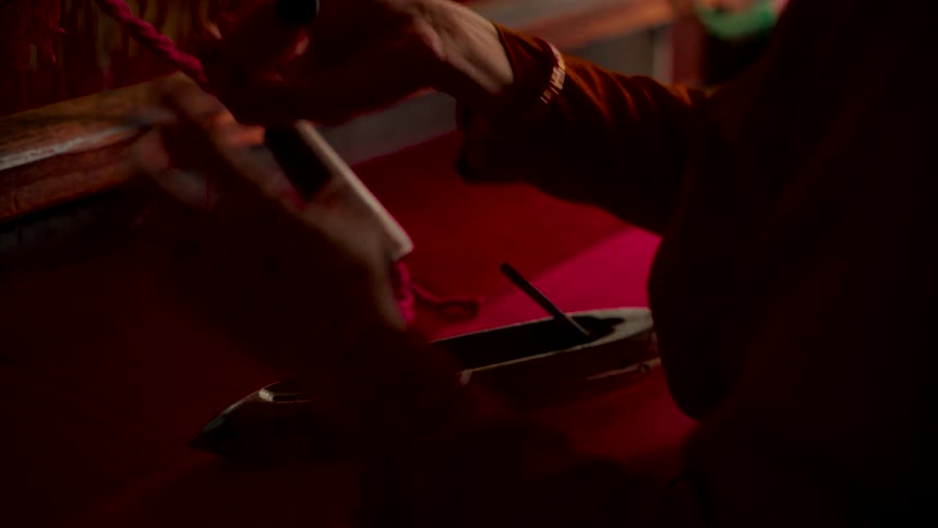 A weaver's hands carefully thread pink yarn on a traditional wooden loom in a dimly lit workshop