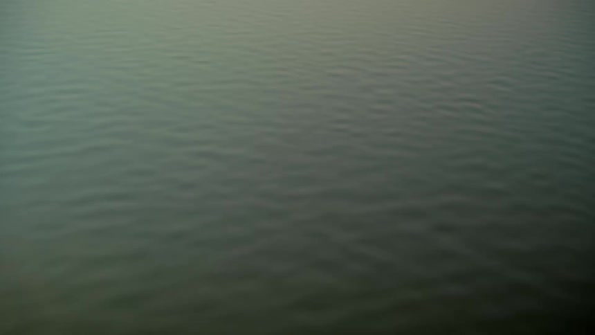 A high-angle, static shot shows the serene surface of a calm body of water with gentle ripples