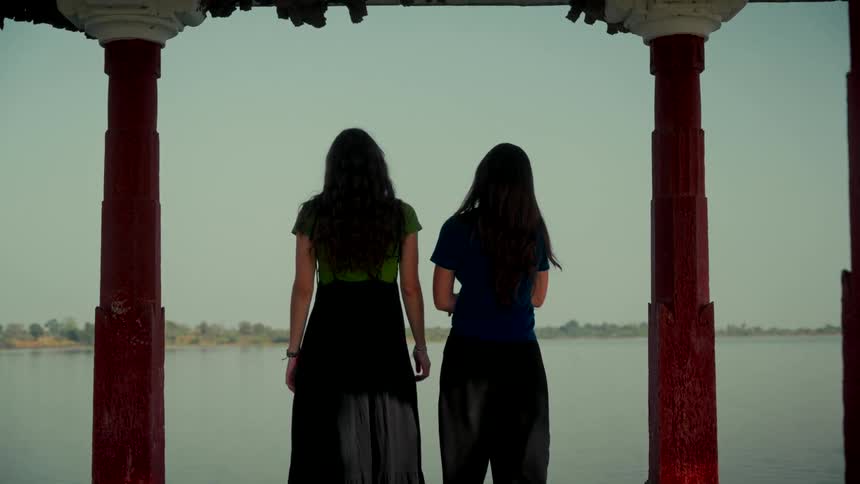 Rear view of two women with long dark hair standing between red pillars and looking out at a vast, calm lake