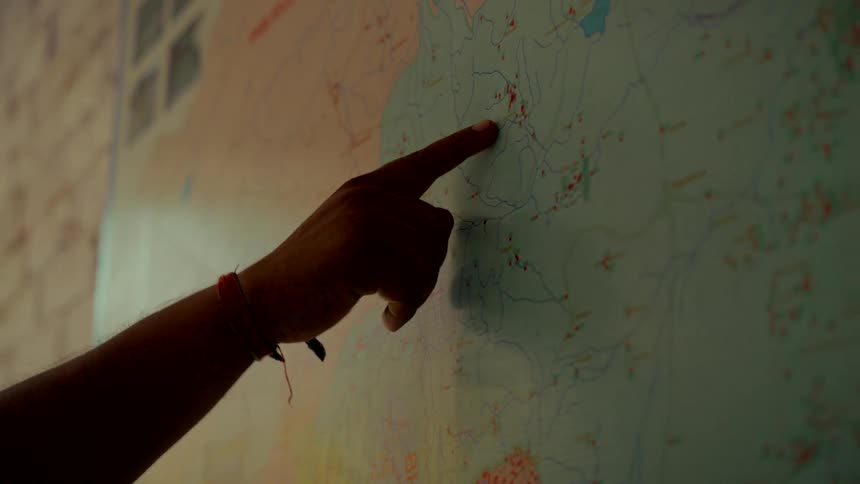 A person's silhouetted hand points to a specific location on a large, detailed wall map