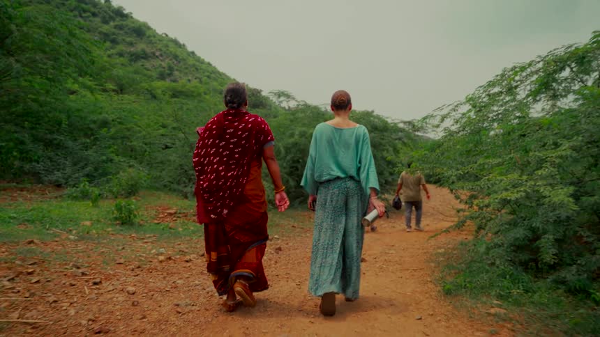A local woman in a traditional sari and a tourist walk together down a dirt path in the lush Indian countryside