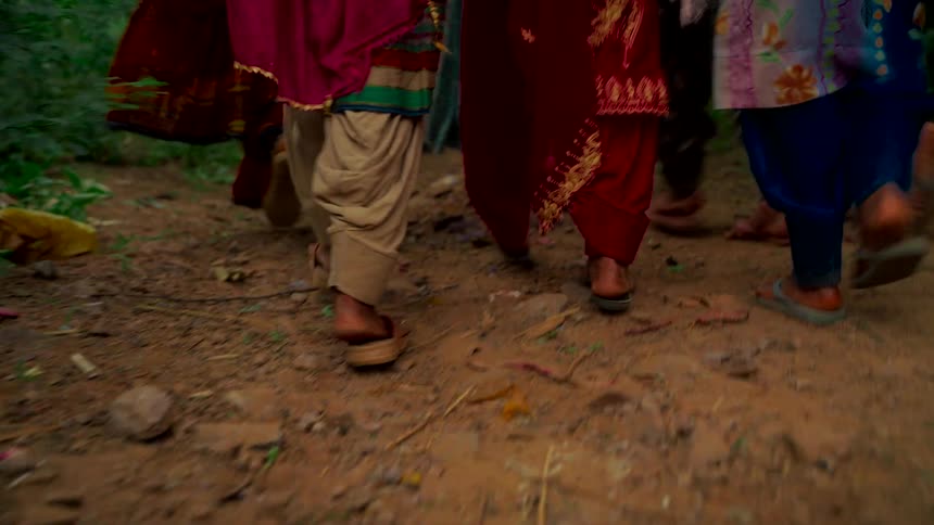 A low-angle, slow-motion shot follows the feet of a group of women in colorful traditional clothes as they walk along a dirt path