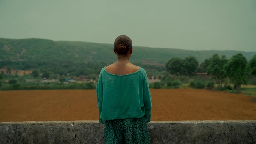 A woman from behind stands on a balcony overlooking a vast rural landscape with tilled fields and green hills on an overcast day