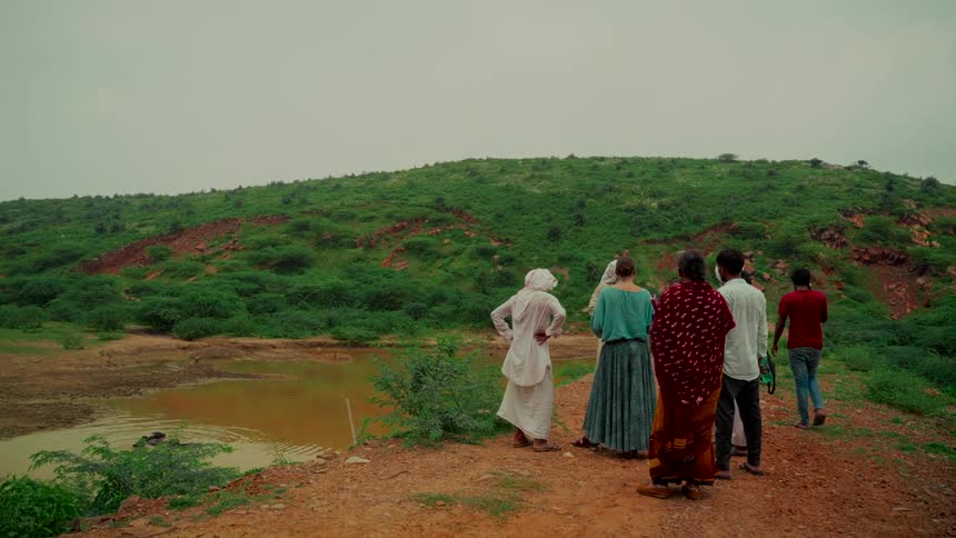 A diverse group of people stand on a dirt path overlooking a verdant green valley and a small pond on an overcast day