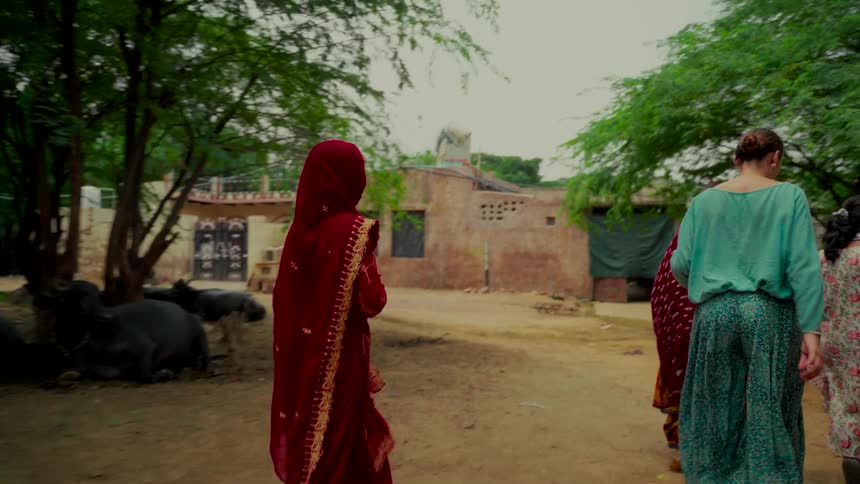 A Western woman and local Indian women walk together through a rural village with livestock nearby