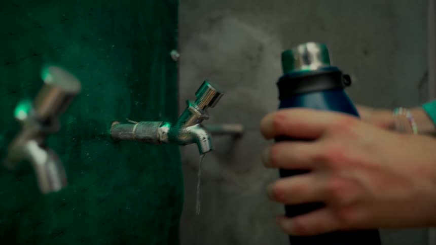 A person in a green shirt fills a reusable water bottle at an outdoor tap and screws the lid on