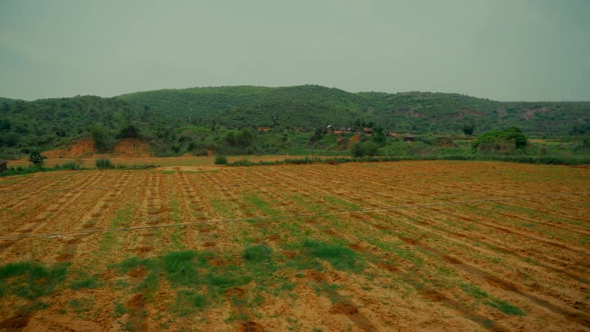 Slow pan of a tilled farm field with irrigation lines at the base of lush green hills under a cloudy sky