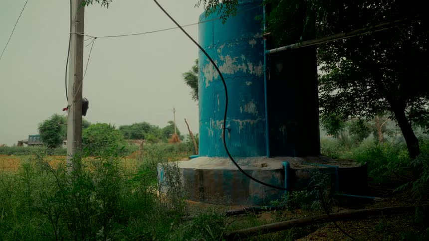A weathered blue water tank sits amongst overgrown bushes and trees in a rural countryside setting