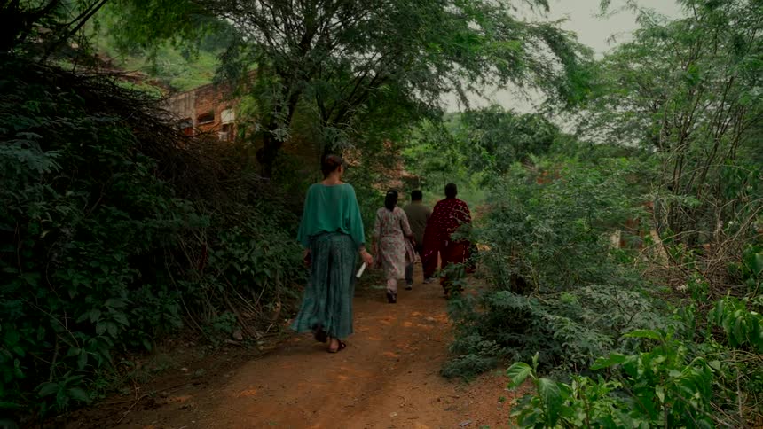 A group of women in colorful clothing walk together down a lush, green dirt path in a rural village