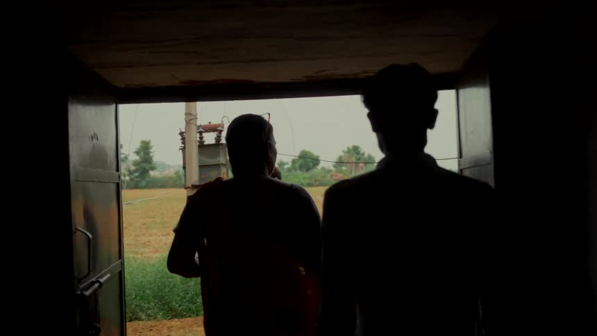 A group of people are silhouetted as they walk out of a dark building into a rural village in India in slow motion