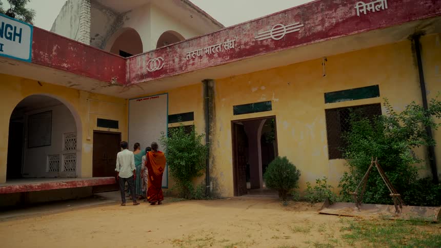 Three villagers stand in the sandy courtyard of a weathered community building in rural India
