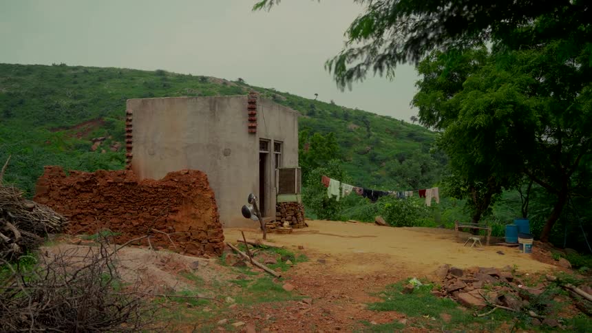 A small, humble home sits in a lush, green hilly landscape with laundry hanging out to dry on an overcast day