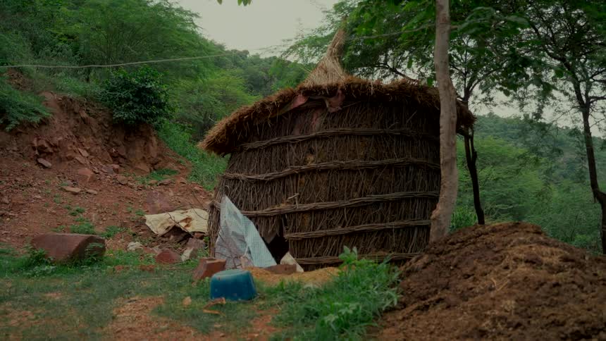 A rustic, handmade hut made of woven branches with a thatched roof sits in a forest clearing at the base of a hill