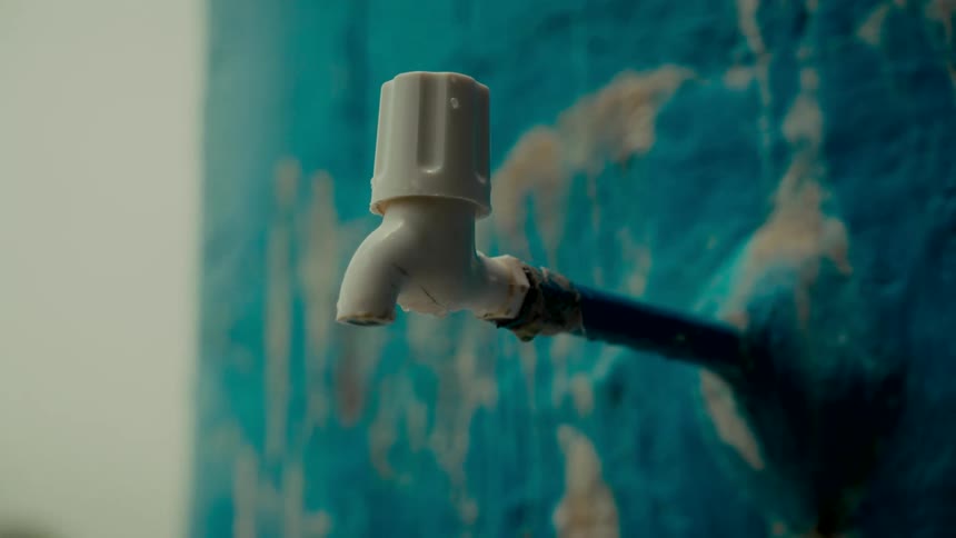 A close-up of a white plastic tap dripping water slowly against a rough, vibrant blue wall