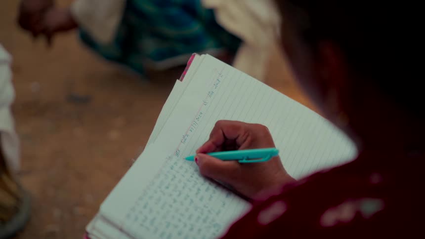 Over the shoulder view of an older Indian woman diligently writing notes in a notebook