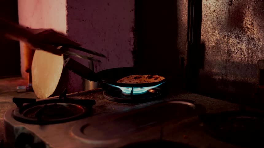 A person flips a traditional homemade Indian flatbread in a pan on a rustic gas stove in a dimly lit kitchen