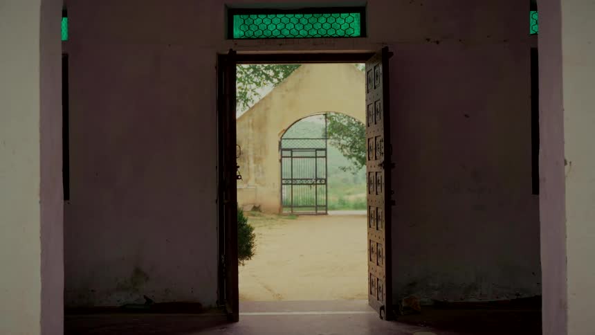 An open wooden door with a green transom window frames a view of a sunlit courtyard and arched gateway