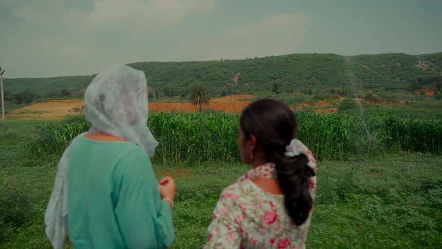 Two Indian women stand together, looking out over a vibrant green crop field being watered by sprinklers in the rural countryside