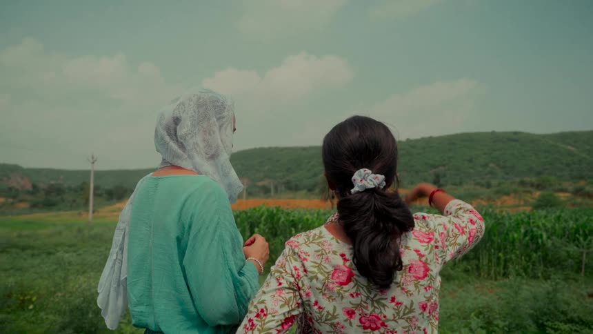 Two Indian women stand side-by-side as they look out at a vast green crop field being watered by sprinklers