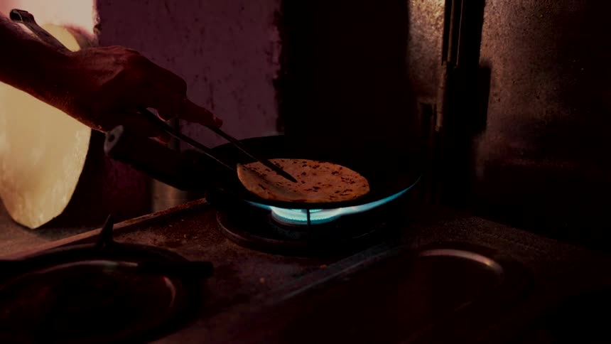A person slowly cooks a traditional Indian flatbread over an open gas flame in a dimly lit, rustic kitchen