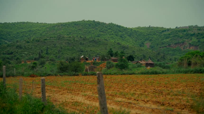 A tranquil scene of a small rural village and plowed farmland at the foot of a lush green mountain