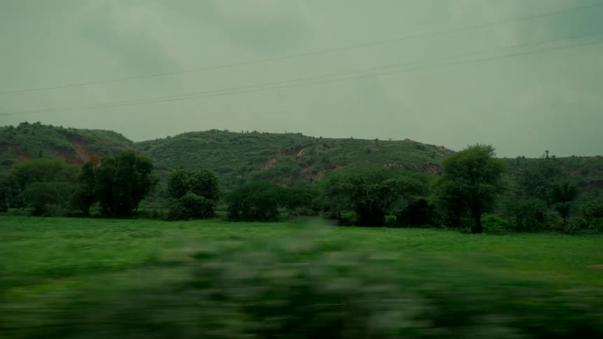 A tracking shot from a moving vehicle reveals the lush green countryside and rolling hills on an overcast day